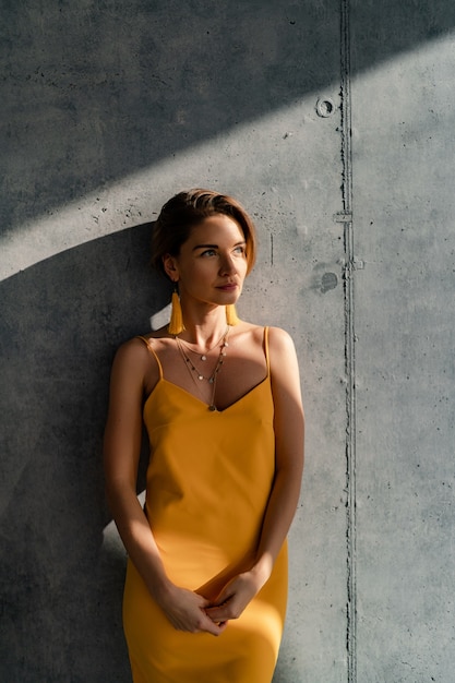 Mujer en vestido amarillo de verano con peinado corto en el interior de la habitación muro de hormigón posando