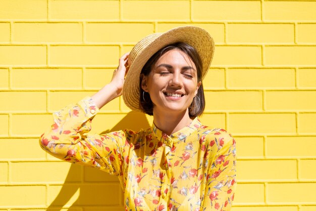 Mujer en vestido amarillo de verano y sombrero en la pared de ladrillo amarillo tranquila y positiva, disfruta de los días soleados de verano