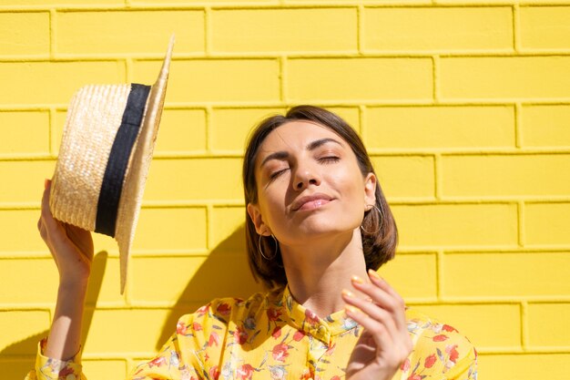 Mujer en vestido amarillo de verano y sombrero en la pared de ladrillo amarillo tranquila y positiva, disfruta de los días soleados de verano