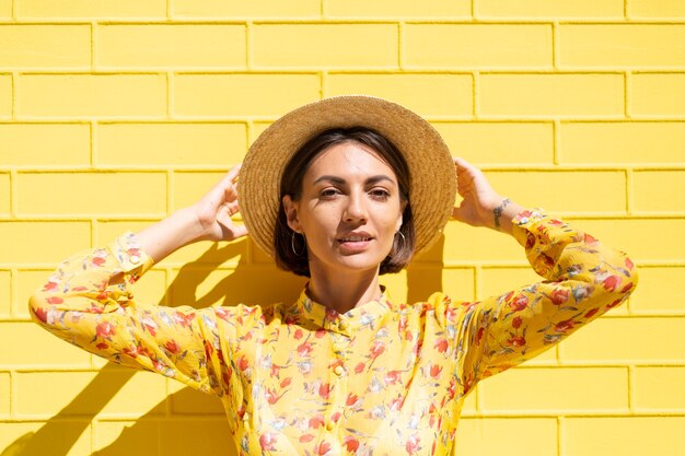 Mujer en vestido amarillo de verano y sombrero en la pared de ladrillo amarillo tranquila y positiva