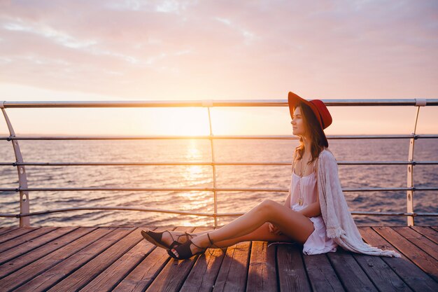 Mujer con vestido blanco sentada junto al mar al amanecer en un estado de ánimo romántico con sombrero rojo