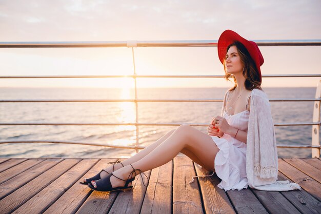 Mujer con vestido blanco sentada junto al mar al amanecer en un estado de ánimo romántico con sombrero rojo