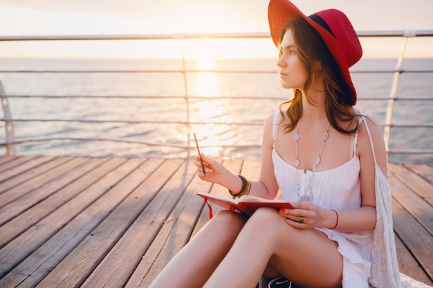 Mujer con vestido blanco sentada junto al mar al amanecer pensando y tomando notas en el libro diario en un estado de ánimo romántico con sombrero rojo