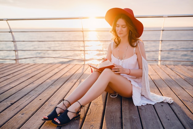 Mujer con vestido blanco sentada junto al mar al amanecer pensando y tomando notas en el libro diario en un estado de ánimo romántico con sombrero rojo