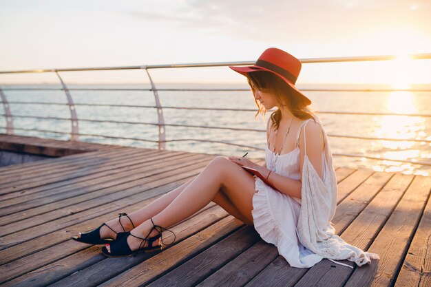 Mujer con vestido blanco sentada junto al mar al amanecer pensando y tomando notas en el libro diario en un estado de ánimo romántico con sombrero rojo