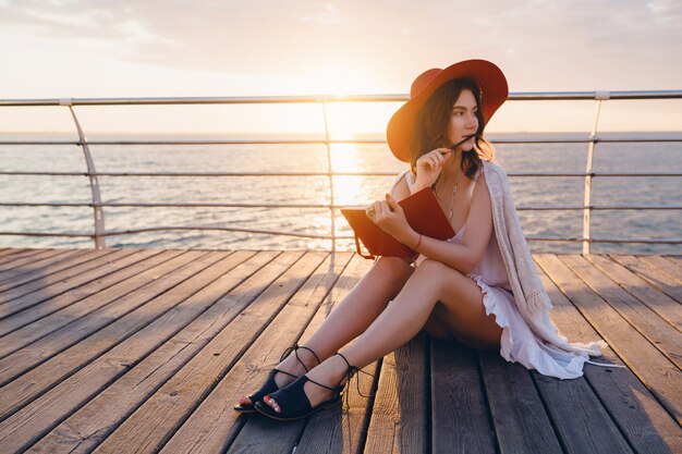 Mujer con vestido blanco sentada junto al mar al amanecer pensando y tomando notas en el libro diario en un estado de ánimo romántico con sombrero rojo