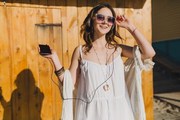 Mujer en vestido blanco de verano escuchando música con auriculares bailando y divirtiéndose