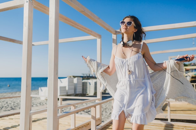 Mujer en vestido blanco de verano escuchando música con auriculares bailando y divirtiéndose