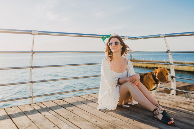 Mujer en vestido blanco de verano jugando con perro junto al mar
