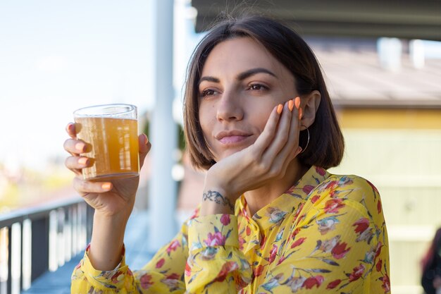 Mujer en vestido en la cafetería de verano disfrutando de un vaso de cerveza kombucha fresca