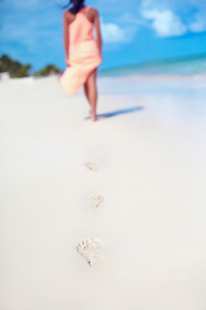 Mujer en vestido colorido caminando en la playa del océano dejando huellas en la arena