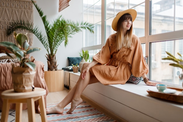 Mujer en vestido de lino relajarse en casa, mirando a la ventana