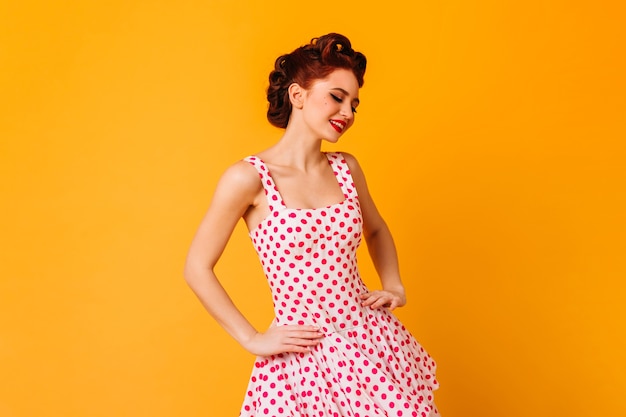 Mujer en vestido de lunares posando con sonrisa. Chica pinup romántica riendo en el espacio amarillo.