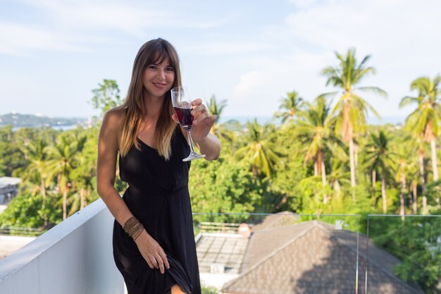 Mujer en vestido de noche negro con copa de vino en el balcón tropical