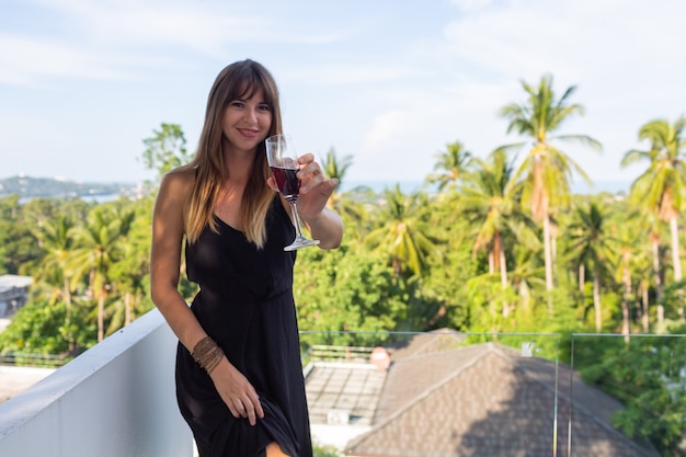 Mujer en vestido de noche negro con copa de vino en el balcón tropical