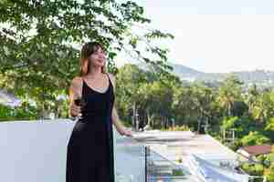 Foto gratuita mujer en vestido de noche negro con copa de vino en el balcón tropical