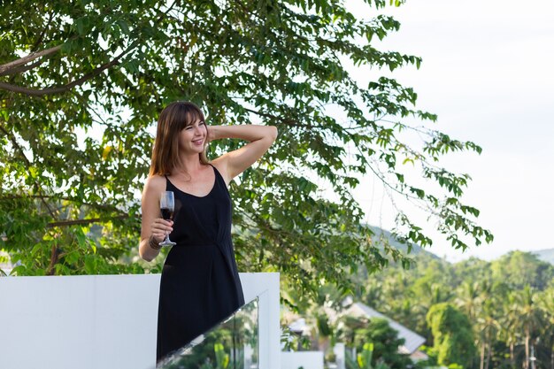 Mujer en vestido de noche negro con copa de vino en el balcón tropical