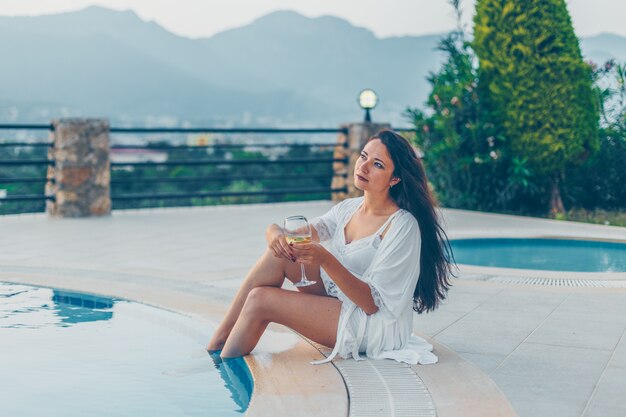 Mujer en vestido de noche sentado en el borde de la piscina y sosteniendo champán en casa durante el día