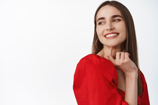 Mujer en vestido rojo romántico, sonriendo y mirando a la izquierda con una sonrisa feliz y complacida, de pie sobre blanco