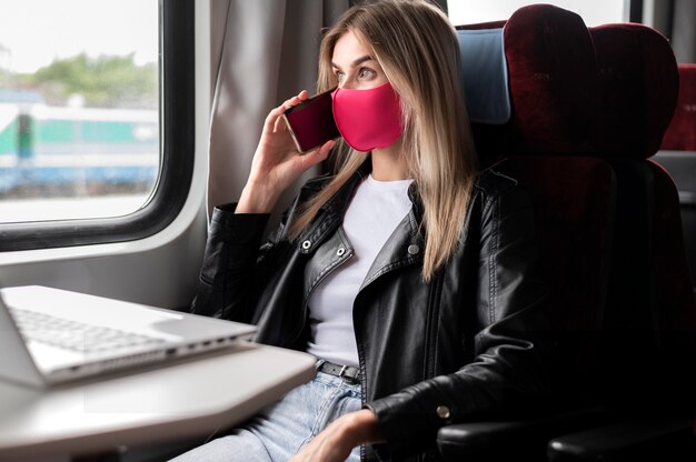 Mujer viajando en tren y hablando por teléfono mientras usa una máscara médica y trabaja en una computadora portátil