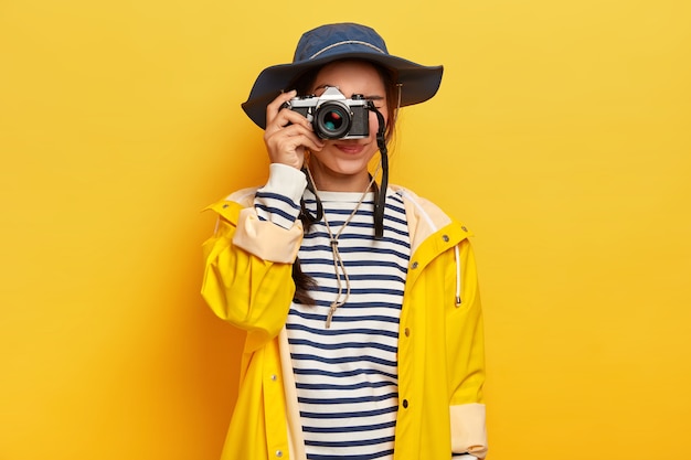 Mujer viajera hace fotos memorables durante el viaje, sostiene una cámara retro, toma imágenes de un hermoso paisaje o lugar, vestida con un jersey de rayas, un impermeable y un sombrero, aislado sobre una pared amarilla