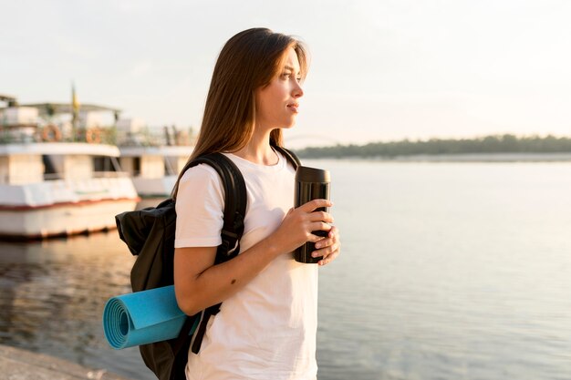 Mujer viajera con mochila sosteniendo termo y admirando el río