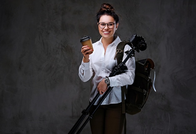 La mujer viajera sostiene una cámara fotográfica digital y café para llevar sobre fondo gris.