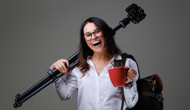 La mujer viajera sostiene una cámara de fotos digital y una taza de café roja sobre un fondo gris.