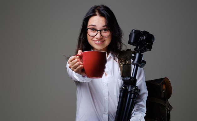 La mujer viajera sostiene una cámara de fotos digital y una taza de café roja sobre un fondo gris.