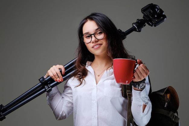 La mujer viajera sostiene una cámara de fotos digital y una taza de café roja sobre un fondo gris.