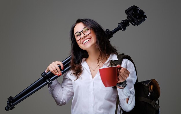 La mujer viajera sostiene una cámara de fotos digital y una taza de café roja sobre un fondo gris.