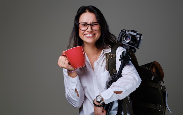 La mujer viajera sostiene una cámara de fotos digital y una taza de café roja sobre un fondo gris.