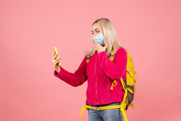 Mujer de viajero de vista frontal con mochila amarilla con máscara tomando una foto con su teléfono