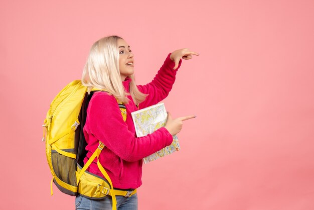 Mujer de viajero de vista frontal con mochila amarilla sosteniendo el mapa en la pared rosa