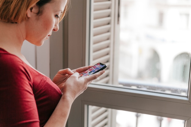 Mujer viendo el teléfono inteligente cerca de la ventana