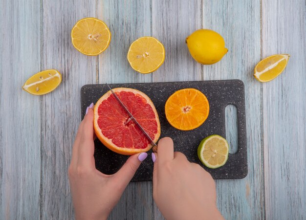 Mujer vista superior corta gajos de pomelo con naranja lima y limón en una tabla de cortar sobre un fondo gris