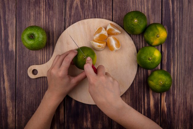 Mujer vista superior corta mandarinas verdes en una tabla de cortar en una pared de madera