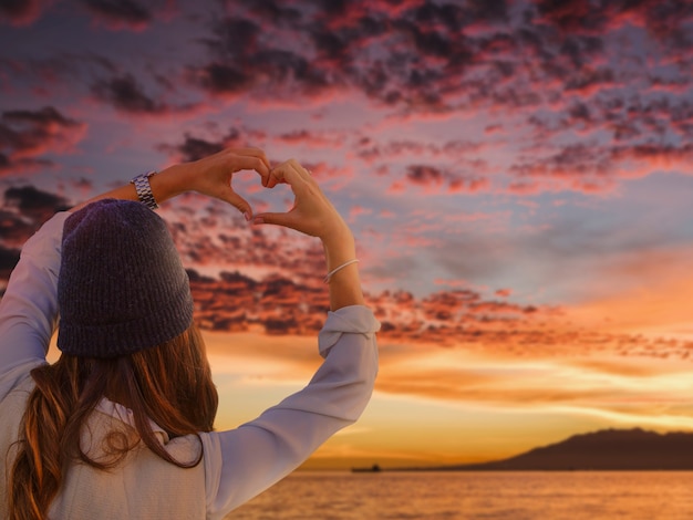 Mujer de vista trasera haciendo manos en forma de corazón en una puesta de sol