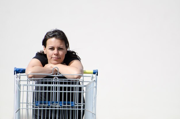 Mujer vistiendo una camisa negra apoyado en un carrito de compras con un fondo blanco.