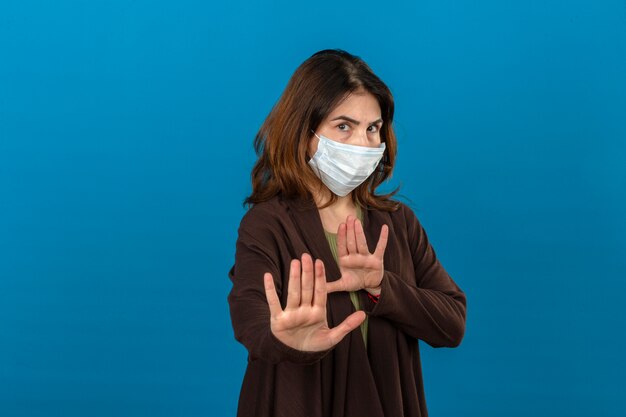 Mujer vistiendo cardigan marrón en máscara protectora médica sosteniendo sus manos diciendo que no se acerque sobre la pared azul aislada