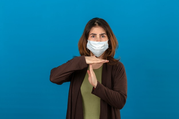 Foto gratuita mujer vistiendo chaqueta marrón en máscara protectora médica mirando cansado haciendo gesto de tiempo con las manos de pie sobre la pared azul aislada