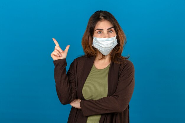 Mujer vistiendo chaqueta marrón en máscara protectora médica mirando seguro apuntando hacia el lado con el dedo parado sobre la pared azul aislada