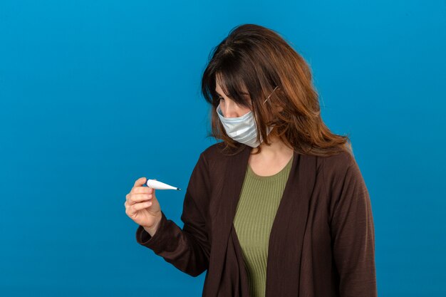 Mujer vistiendo chaqueta marrón en máscara protectora médica mirando el termómetro digital mirando nervioso sobre la pared azul aislada