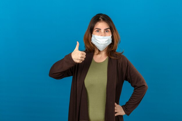 Mujer vistiendo chaqueta marrón en máscara protectora médica mostrando el pulgar hacia arriba sobre la pared azul aislada
