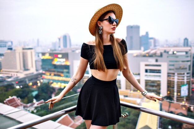 Mujer vistiendo crop top y minifalda, sombrero de paja y accesorios de moda, posando en la azotea del hotel de lujo en la gran ciudad moderna