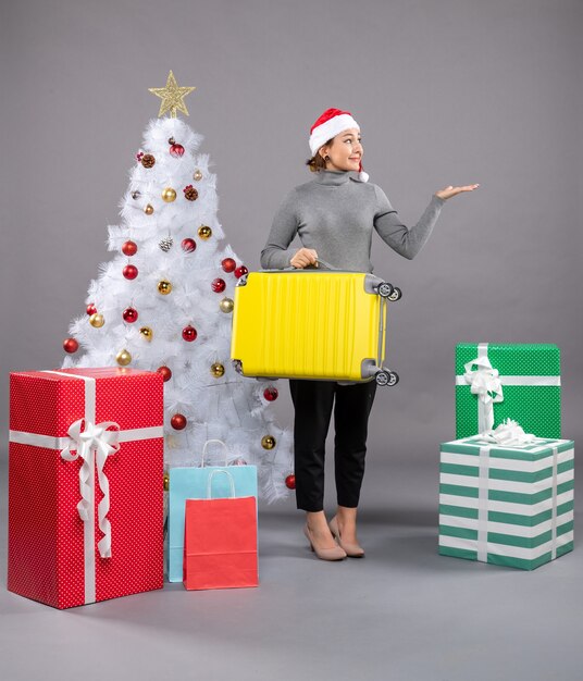 Mujer vistiendo gorro de Papá Noel con equipaje junto al árbol de Navidad