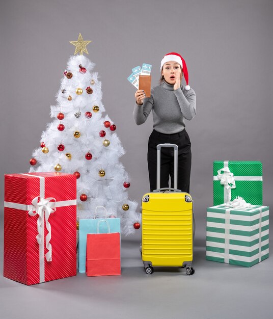 Mujer vistiendo gorro de Papá Noel con equipaje junto al árbol de Navidad