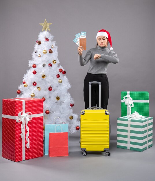 Mujer vistiendo gorro de Papá Noel con equipaje junto al árbol de Navidad