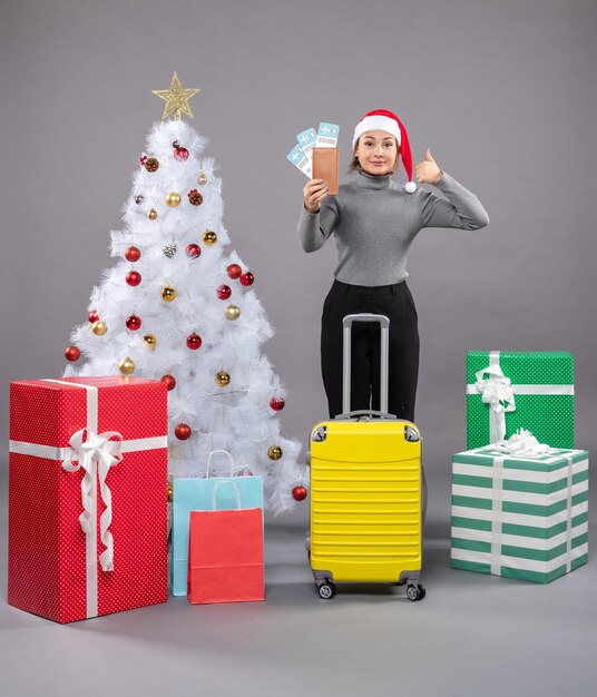 Mujer vistiendo gorro de Papá Noel con equipaje junto al árbol de Navidad