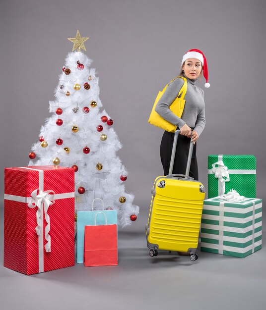 Mujer vistiendo gorro de Papá Noel con equipaje junto al árbol de Navidad